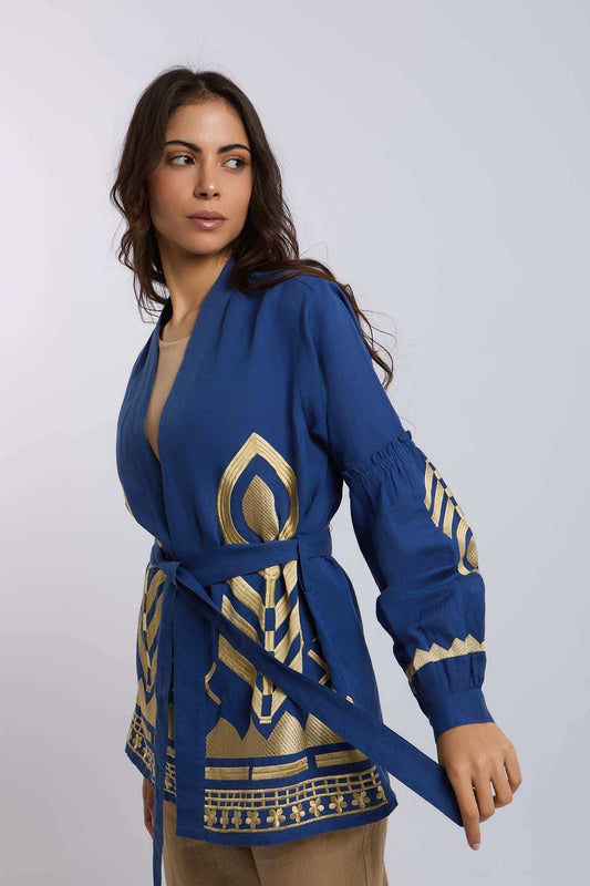 Woman wearing blue belted jacket with gold geometric embroidery, standing against light background